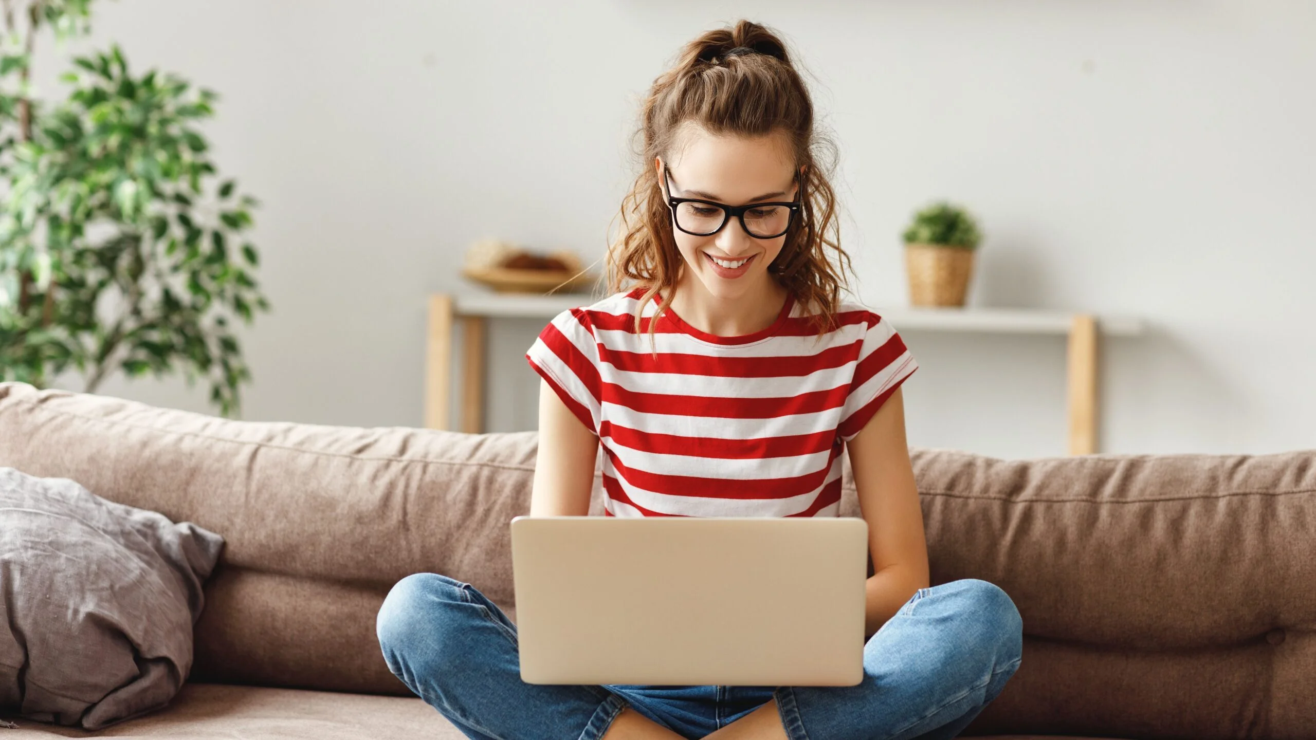 Garota usando óculos e blusa listrada mexendo em seu notebook sentada no sofá de sua casa.